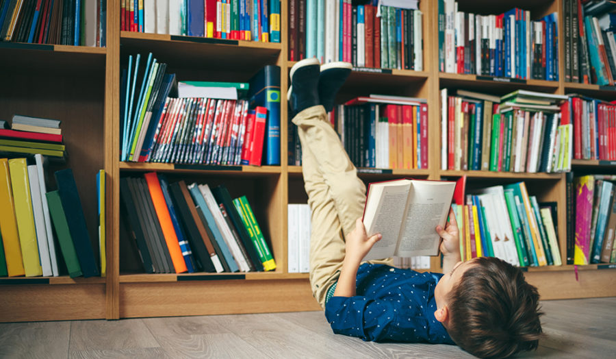 Boy laying on the floor with the feet up, reading a book against multi colored bookshelf in library. Education, Knowledge, Bookstore, Lecture. Pupil holds a book in his hands.